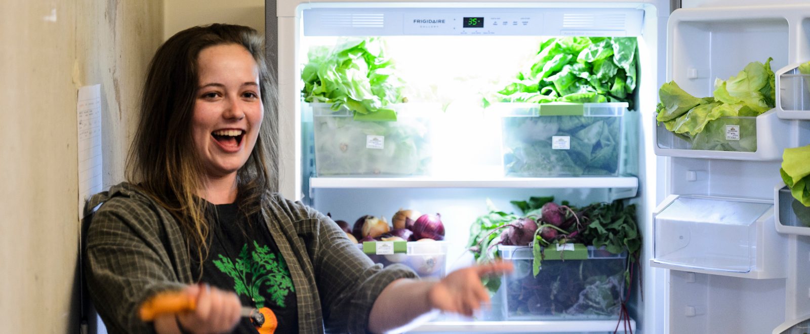 Hannah DePorter stands in front of the new refrigerator, door open displaying a bounty of fresh greens, onions, beets.