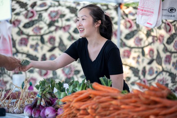 Surrounded by carrots, onions and garlic, Naomi Moua helps a customer at her family’s booth at a farmers’ market.